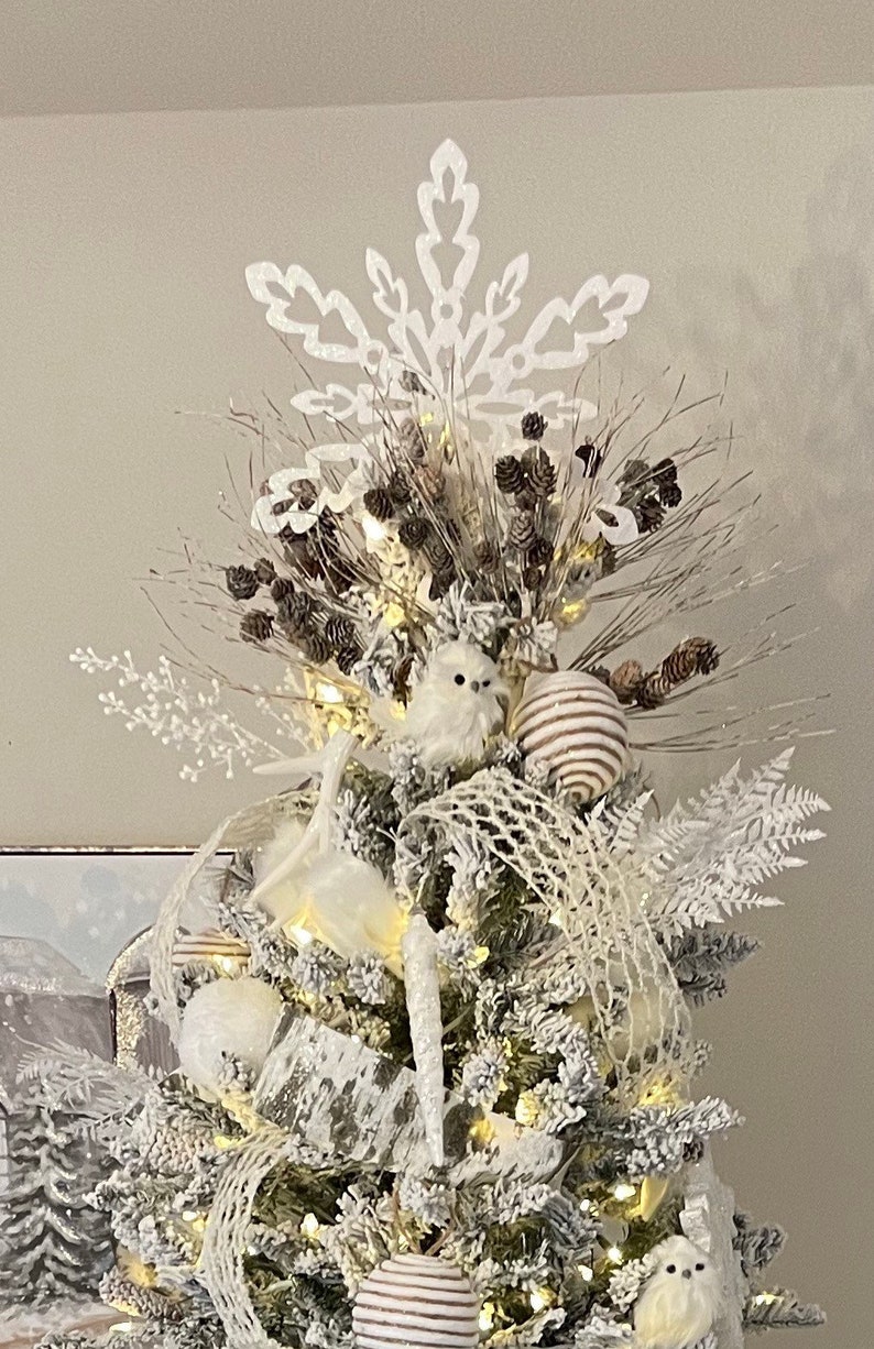 May include: A white flocked Christmas tree decorated with white and brown ornaments, a white snowflake tree topper, and white owls. The tree is lit with white fairy lights.