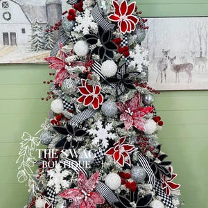 May include: A decorated Christmas tree with red, black, and white ornaments. The tree features red poinsettias, black and white checkered ribbon, silver ornaments, and snowflake decorations. The tree is in front of a painting of a barn and a painting of deer.