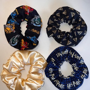May include: Four scrunchies in various designs. One black scrunchie features gold symbols, another has a black background with colorful crests, a third is a shiny gold satin, and the last is blue with white text.