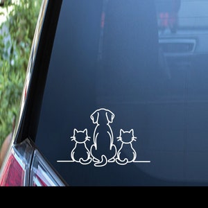May include: White vinyl decal of a dog and two cats sitting in a row. The dog is in the middle and the cats are on either side. The decal is on the back window of a car.