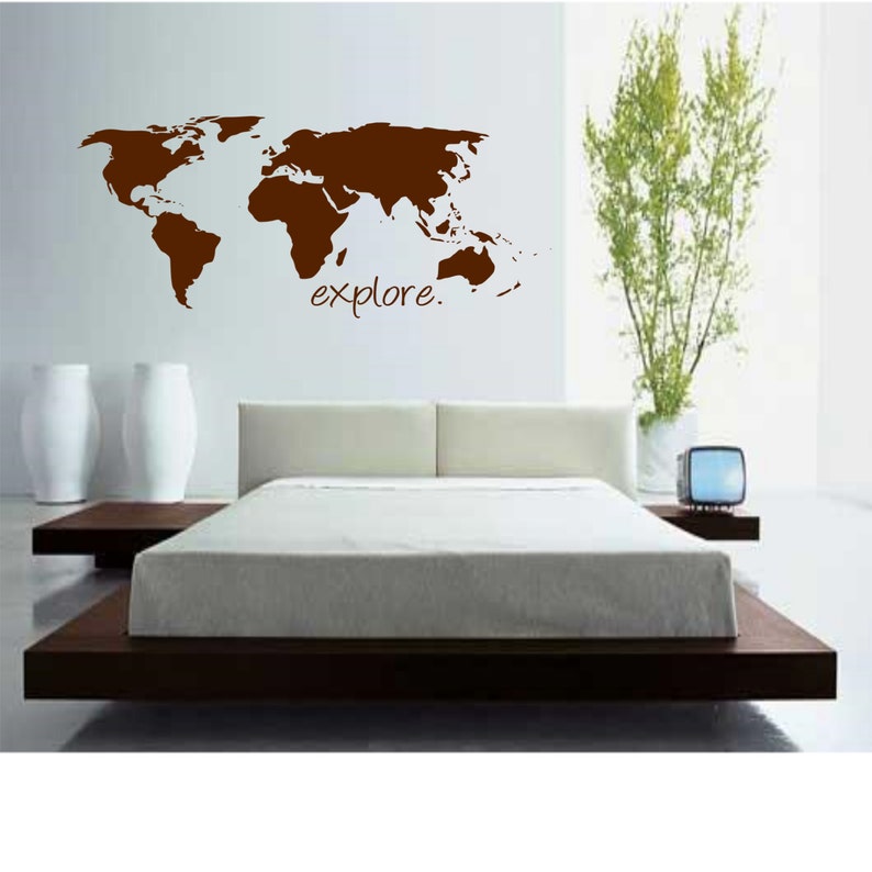 K&ouml;nnte beinhalten: Ein brauner Wandaufkleber mit einer Weltkarte und dem Wort "explore" darunter. Der Aufkleber befindet sich an einer wei&szlig;en Wand &uuml;ber einem Bett mit einem braunen Kopfteil und einer wei&szlig;en Bettdecke.