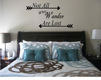 May include: A bedroom with a black headboard and a bed with a blue and brown patterned duvet. The wall above the headboard has a black vinyl decal with the text "Not All Who Wander Are Lost" with arrows on either side.