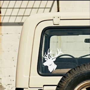 May include: A white vinyl decal of a deer head with large antlers is affixed to the rear window of a cream-colored vehicle. The decal is a silhouette, showcasing the deer's profile. The vehicle's tire is visible.