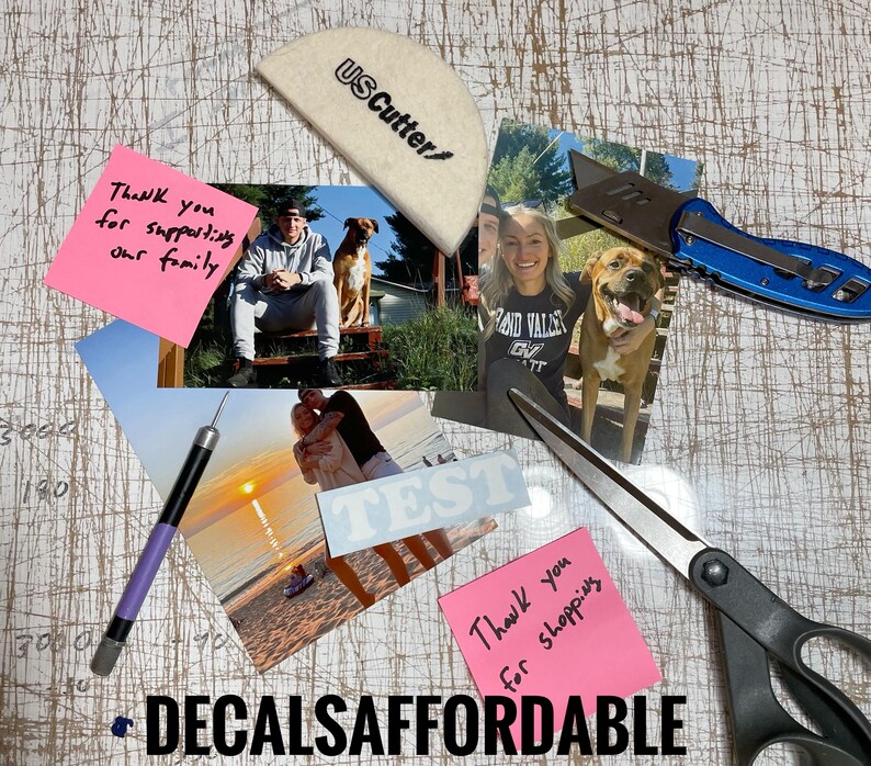 May include: A close-up of a table with various items, including a pink sticky note with the text "Thank you for supporting our family", a white cutting mat with the text "USCutter", a pair of scissors, a utility knife, and several photographs.