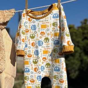 May include: White baby onesie with a pattern of colorful cartoon monsters. The onesie has a yellow trim around the neck and cuffs.