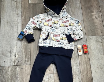 May include: A white hooded sweatshirt with a navy blue lining and a pattern of colorful trucks. The sweatshirt is paired with navy blue sweatpants.