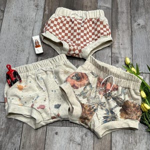 May include: Three pairs of toddler shorts in beige with different patterns. One pair has a checkered pattern in brown and beige, another has a floral pattern in shades of brown, orange, and blue, and the last pair has a splatter paint design in shades of brown, orange, and blue.