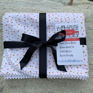 May include: A white organic flannel baby blanket with a pink, gold, and gray polka dot pattern. The blanket is tied with a black ribbon and has a white pom-pom trim. The tag reads "Organic Flannel Blanket Sewn for you by M.L. Machine Wash Cold Tumble Dry Low Remove Promptly www.SewnForYouByML.etsy.com"