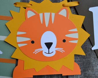 May include: Orange and yellow lion head banner with a white face and black nose. The lion has stripes on its face and whiskers.