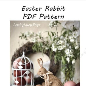 May include: A decorative Easter rabbit figurine, approximately 4.7 inches (12 cm) tall, with a cream-colored bow, sits on a small wooden cart. The image includes a white wire cage with red eggs, and the text "Easter Rabbit PDF Pattern".