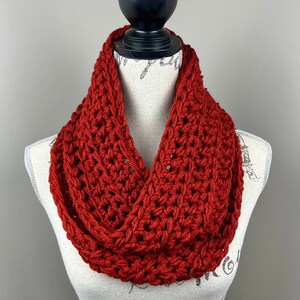 May include: A rust-colored, chunky knit infinity scarf displayed on a mannequin. The scarf is made of thick yarn, with a visible stitch pattern. The warm, earthy tone of the scarf suggests a cozy, handmade accessory, perfect for cooler weather.