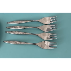 May include: Four silver forks with textured handles and three tines each. The forks are arranged on a teal background. The handles have a geometric pattern. The forks are made of stainless steel.