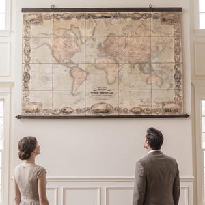A large empty wall is transformed with a stunning oversized world map canvas. A happily married couple stands together, admiring their beautiful wedding gift with smiles and excitement.