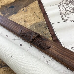 May include: A close-up of a wooden frame with an ornate carved design. The frame is holding a rolled-up map with a compass rose and markings in degrees. The map is titled "Plan de Monaco".