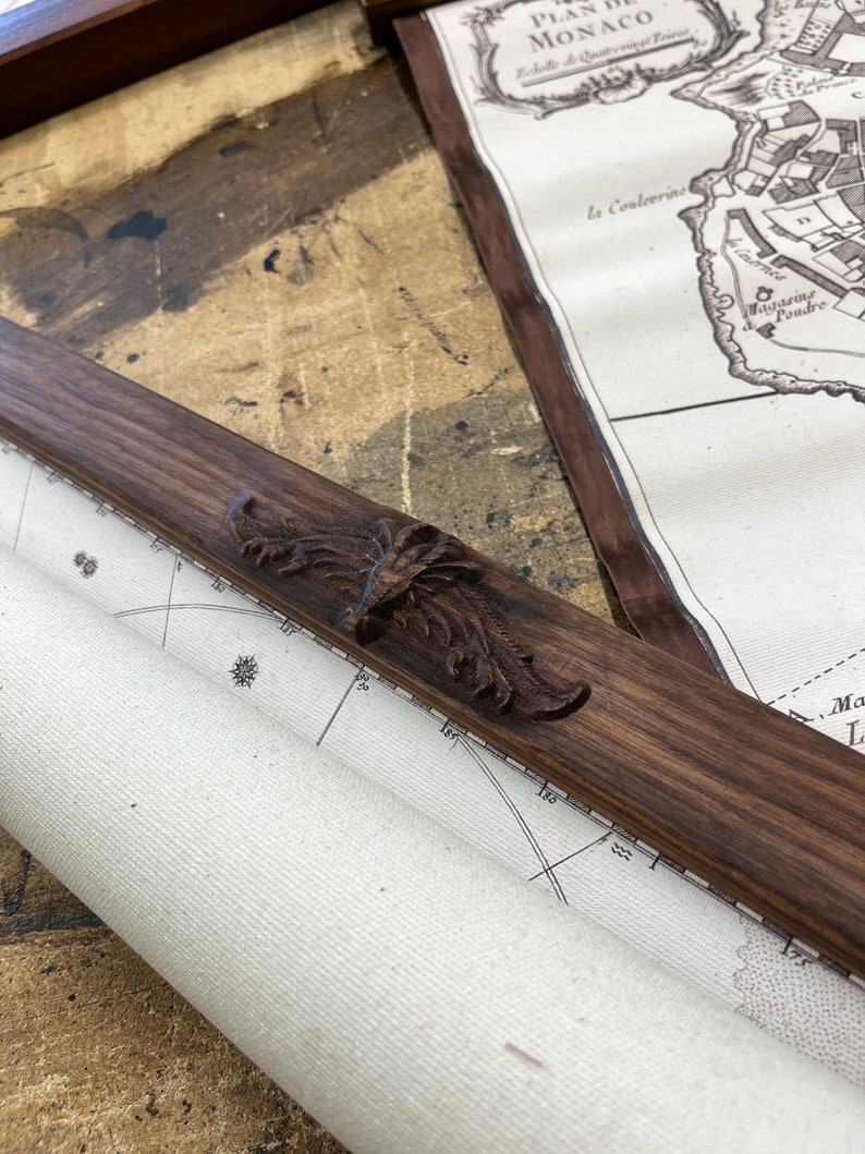 May include: A close-up of a wooden frame with an ornate carved design. The frame is holding a rolled-up map with a compass rose and markings in degrees. The map is titled "Plan de Monaco".