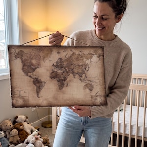 May include: A rectangular wall hanging featuring a vintage-style world map printed in sepia tones on a textured surface. The map displays continents and oceans with a grid overlay and is suspended by a thin, light brown twine cord attached to the top.