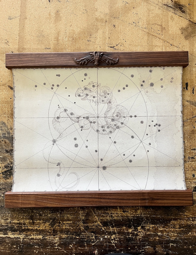 May include: A vintage-style map of a constellation, framed in a dark wood frame with a decorative carved wooden top. The map shows a constellation of stars in the shape of a dragon.