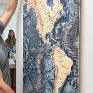 May include: A vertical wall map of the Americas featuring detailed bathymetric ocean floor topography in shades of deep blue and tan landmasses. The map is secured between two black wooden rods at the top and bottom with a black hanging cord.
