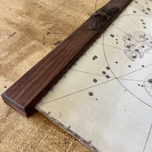 May include: A close-up of a wooden map hanger with a decorative scroll design. The hanger is made of dark wood and has a natural finish. The hanger is attached to a map with a white background and black lines.
