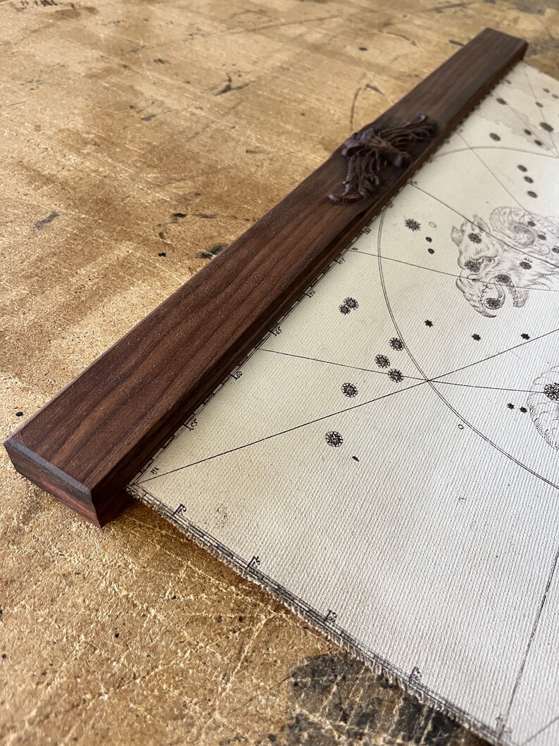 May include: A close-up of a wooden map hanger with a decorative scroll design. The hanger is made of dark wood and has a natural finish. The hanger is attached to a map with a white background and black lines.