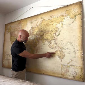 May include: A large, vintage-style wall map of the world featuring a sepia-toned color palette. The map displays continents, countries, and oceans with detailed cartographic markings, suspended from a wooden rod with a thin string for hanging.
