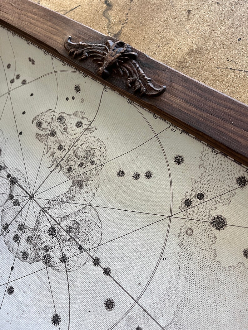 May include: A vintage celestial map framed in dark wood with a decorative metal corner piece. The map features a detailed illustration of a constellation, possibly Draco, with a serpentine shape and numerous stars.