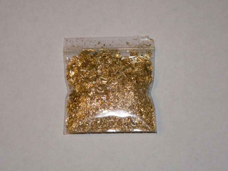 May include: A clear plastic bag filled with gold leaf flakes. The flakes are small, irregular shapes, and reflect light, creating a shimmering effect. The bag is sealed at the top and sits on a plain, light-colored surface.