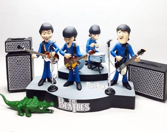 Puede incluir: Un conjunto de figuras que representan a los miembros de la banda The Beatles con trajes azules, tocando instrumentos en un escenario. Incluye amplificadores, una batería y una figura de cocodrilo verde. El logotipo de The Beatles es visible.