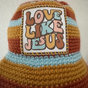 May include: A crocheted bucket hat with a patch that reads "LOVE LIKE JESUS." The hat features horizontal stripes in shades of brown, blue, and yellow. The patch is square with a white background and the text is in various colors.