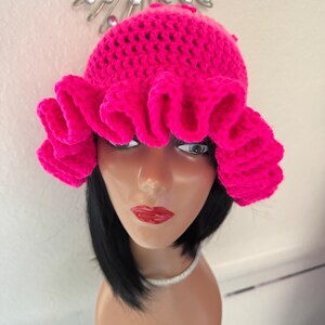 May include: A bright pink crochet hat with a ruffled brim. The hat is made of a soft, textured yarn and has a rounded crown. The vivid colour makes it a fun and stylish accessory.