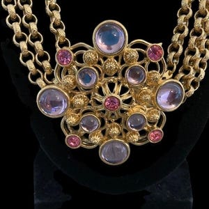 May include: Gold-toned necklace with a multi-strand chain and a large pendant. The pendant features a floral design with various sizes of pink and lavender-colored gemstones. The necklace is displayed against a black background.