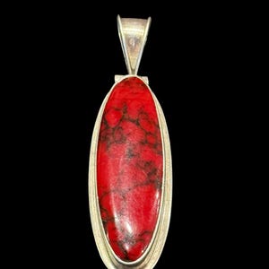 May include: A silver pendant with an oval, red stone. The stone has dark, web-like patterns. The pendant has a silver bail at the top for a chain. The background is black.