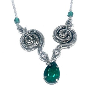 Pode incluir: Colar prateado com dois desenhos de serpentes enroladas com detalhes intrincados. O colar tem um pingente de pedra preciosa verde esmeralda em forma de gota e pequenas contas verdes. A corrente é delicada e sobre um fundo branco.