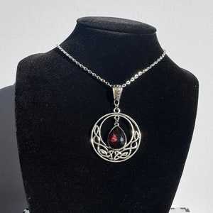 Pode incluir: Um colar de prata com um pingente de nó celta com uma gema de cor púrpura escura. O pingente é circular com um design detalhado.