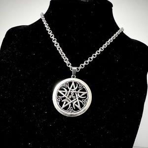 Peut inclure: Collier pendentif argenté avec un motif circulaire orné d'un pentagramme complexe. Le pendentif est suspendu à une chaîne avec un fermoir mousqueton. Le collier est présenté sur un mannequin noir.
