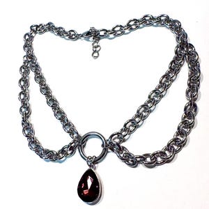 May include: Silver chain necklace with a dark red, teardrop-shaped pendant. The necklace has a double-layered chain design, with a circular ring at the centre. The pendant is a deep burgundy colour, providing a striking contrast to the silver chains.