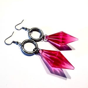 May include: A pair of earrings with a geometric design. Each earring has a bright pink, diamond-shaped pendant hanging from a silver-coloured ring and hook. The earrings are set against a white background.