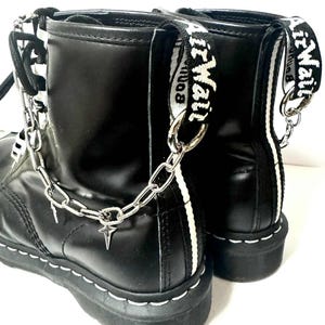 以下が含まれることがあります： 白ステッチと背面に白いストライプが入った黒革ブーツ。ブーツにはシルバーチェーンと星のチャーム、白い「Air Wair」の文字が入ったストラップが付いています。クラシックなスタイルです。