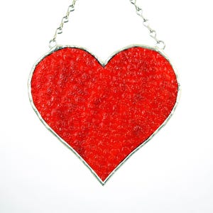 May include: A red heart-shaped stained glass suncatcher with a textured surface. The heart is outlined with a silver-colored metal and hangs from a delicate chain. The suncatcher is a decorative item.