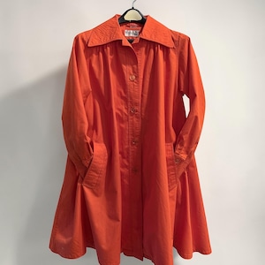 May include: A bright orange, vintage-style coat with a collared neckline and button-down front. The coat features long sleeves, pockets, and a flared skirt. It is hanging on a black hanger against a white background.