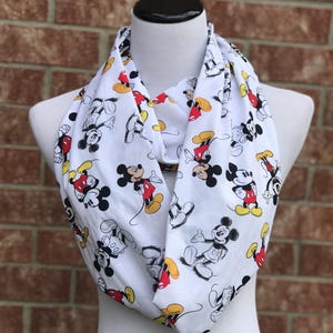 May include: White infinity scarf featuring a repeating pattern of Mickey Mouse in various poses. The scarf has a black, red, yellow, and white color scheme. The scarf is displayed on a mannequin.