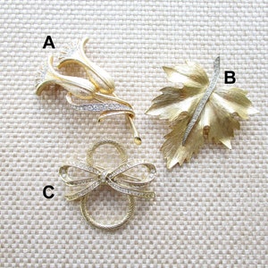 May include: Three gold tone brooches with rhinestones. The first brooch is a cluster of stylized flowers. The second brooch is a leaf with a rhinestone stem. The third brooch is a bow with rhinestones.