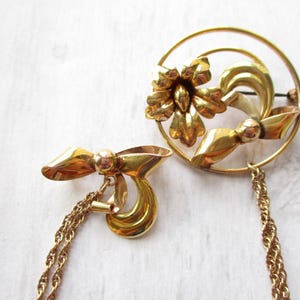 May include: A pair of gold tone brooches with a floral design. One brooch is a circle with a flower and leaves, the other is a bow with a chain.