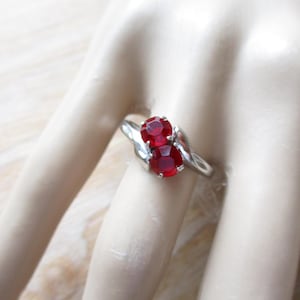 May include: A silver ring featuring two oval-cut, deep red gemstones. The gemstones are set side-by-side, with a swirling silver band that wraps around the finger. The ring is displayed on a pale, neutral background.