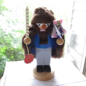 May include: A wooden nutcracker figurine with a brown fur-like head, blue jacket, and gray pants. It holds a white and gold staff and a small mouse figurine with a sword. The base is wood, and a red tag hangs from the staff.