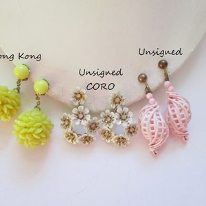 May include: Three pairs of vintage clip-on earrings. The first pair is yellow and features a flower design. The second pair is white and features a flower design. The third pair is pink and features a woven design. The earrings are labeled "Hong Kong", "Unsigned CORO", and "Unsigned".