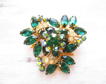 May include: A large, gold-tone brooch with a cluster of emerald green and clear rhinestones. The rhinestones are arranged in a floral pattern, with the emerald green stones forming the petals.