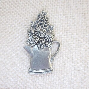 May include: A silver-colored metal brooch in the shape of a watering can overflowing with flowers. The brooch has a detailed design, with visible leaves and flower petals. The background is a light beige woven fabric.