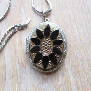 May include: Silver-tone oval locket necklace with a black and gold floral design. The locket has a detailed, textured border and a matching silver chain. The floral design features black petals and a gold center.