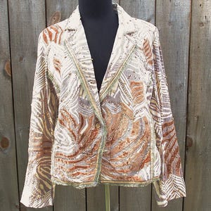 May include: A patterned blazer in shades of cream, brown, and gray, displayed on a mannequin. The jacket features a textured design with a button closure and decorative trim along the edges. The sleeves are long, and the blazer has a classic lapel.